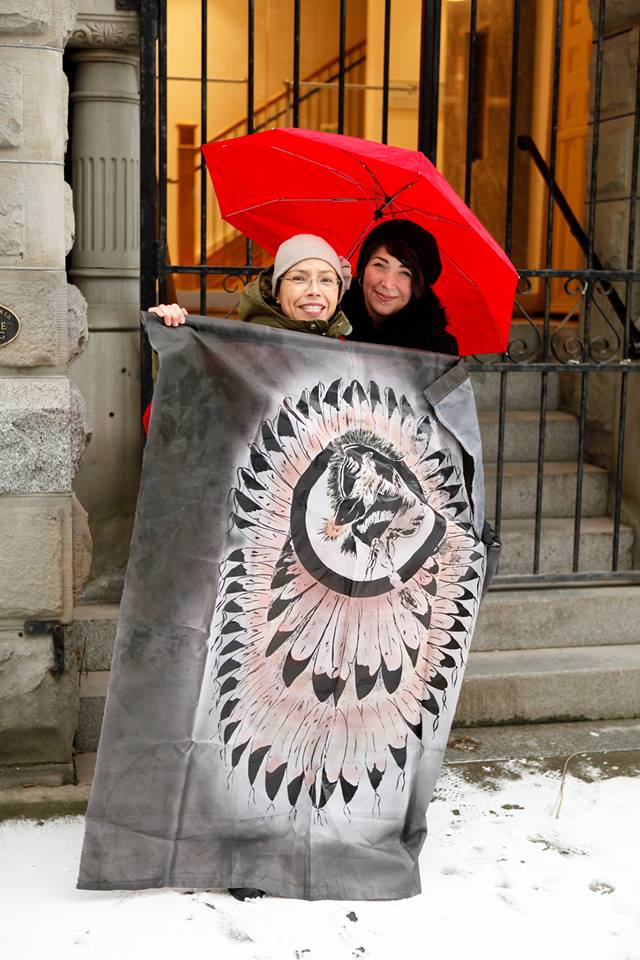 Two people smiling; one holds a red umbrella, the second a blanket with an image of a person in a feather bonnet
